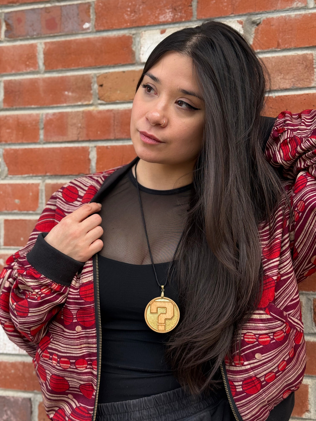 Gorgeous woman wearing a laser cut wooden pendant with a question mark and a red and black patterned jacket with a brick wall background