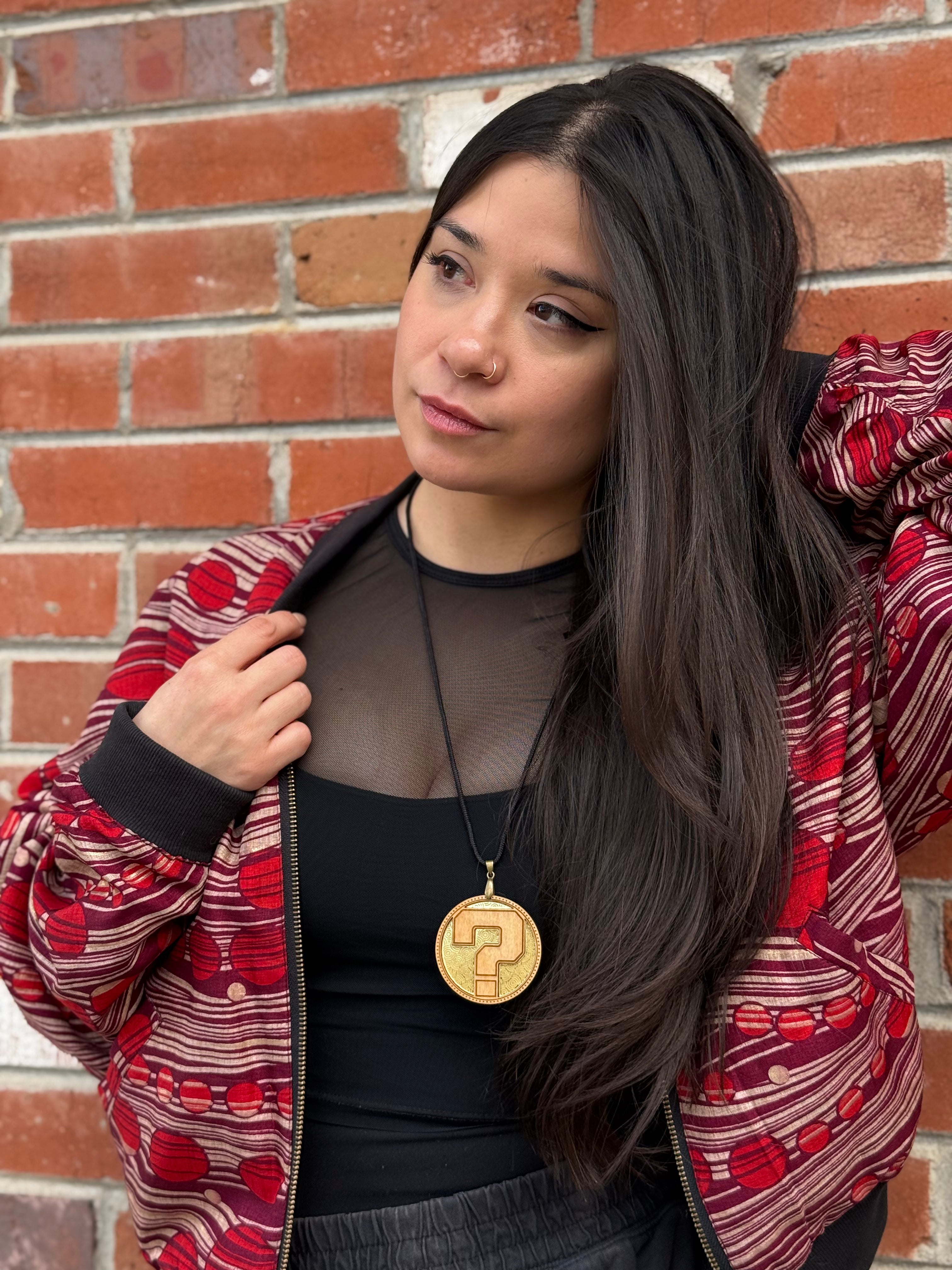 Gorgeous woman wearing a laser cut wooden pendant with a question mark and a red and black patterned jacket with a brick wall background